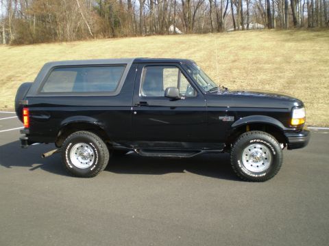1996 Ford Bronco - The side view of a black 1996 Ford Bronco is shown parked on an asphalt surface with a grassy hill and trees in the background. The vehicle…