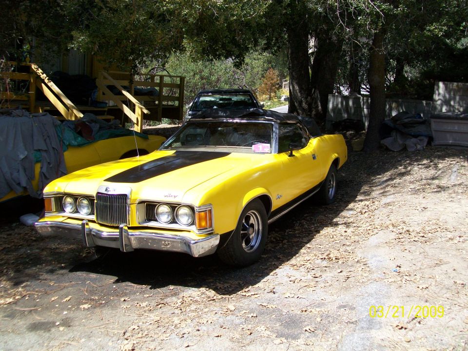 Yellow Mercury Cougar convertible with black top parked on a dirt driveway under trees