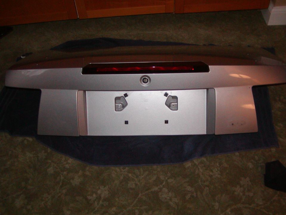 Silver car trunk lid with red reflector and license plate area on a carpet.