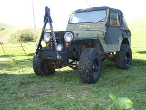 1989 Ford Bronco - A modified olive green Jeep CJ-5 is parked on a grassy hill. The vehicle features large off-road tires, a roll cage, and a soft top.