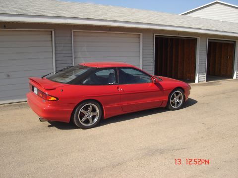 1993 Ford Probe - A red 1993 Ford Probe is parked in front of a garage with two open bays. The car is viewed from the rear three-quarter angle, showcasing it…