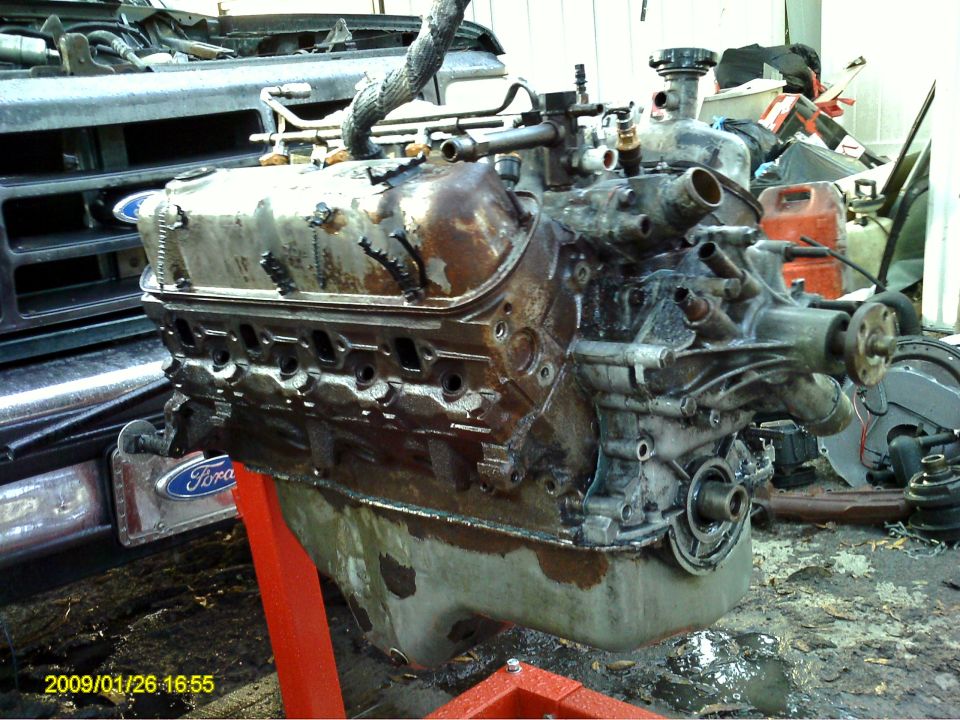 Dirty V8 engine on a red stand, with a Ford emblem visible on a vehicle behind it.