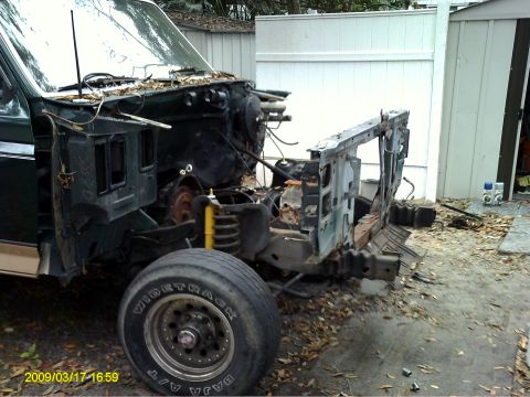 1992 Ford Bronco - The front end of a disassembled dark green vehicle, likely a 1992 Ford Bronco, is shown with the hood and fenders removed, revealing the en…
