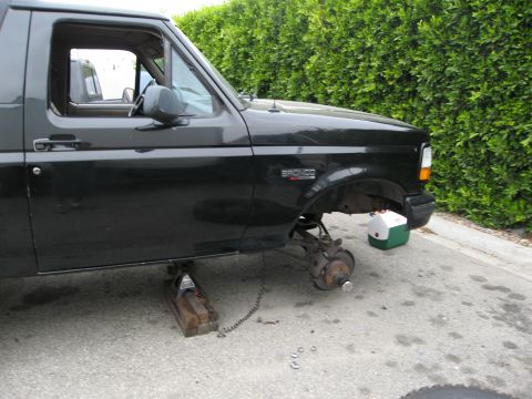 1986 Ford Bronco - The side view of a black Ford Bronco is shown with the front passenger-side wheel removed and the vehicle supported by a jack and wooden bl…