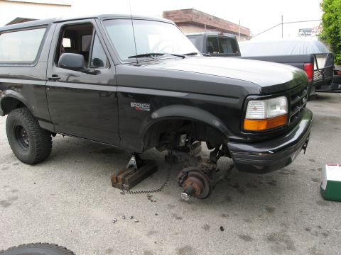 1986 Ford Bronco - A black Ford Bronco is jacked up with its front passenger-side wheel and suspension removed, revealing the brake rotor and hub assembly. Th…