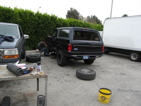 1986 Ford Bronco - A black Ford Bronco is parked on asphalt next to a gray pickup truck, with a person working underneath the Bronco. Several car parts, inclu…