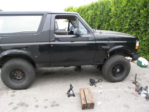 1986 Ford Bronco - The side view of a black Ford Bronco is shown parked on asphalt in front of a green hedge. The vehicle appears to be undergoing maintenance…