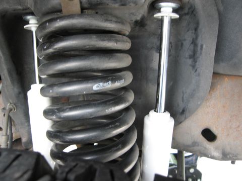 1986 Ford Bronco - A close-up, low-angle view of the front suspension of a vehicle, featuring a large black Eibach coil spring and a white shock absorber. The…