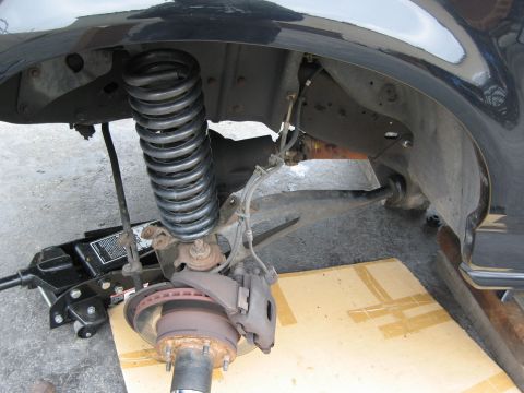 1986 Ford Bronco - A close-up view of the front suspension of a vehicle, showing a large coil spring, brake rotor, caliper, and a hydraulic jack supporting th…