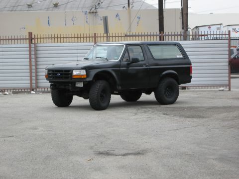 1986 Ford Bronco - A black Ford Bronco is parked on a paved lot in front of a corrugated metal fence and a building. The vehicle has large off-road tires and…