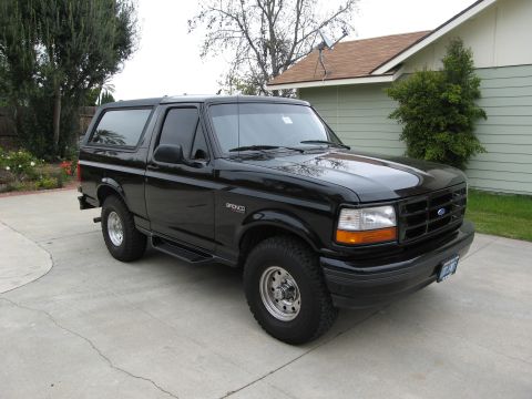 1986 Ford Bronco - A black 1980s Ford Bronco is parked on a concrete driveway in front of a light green house. The SUV has tinted windows and chrome wheels.