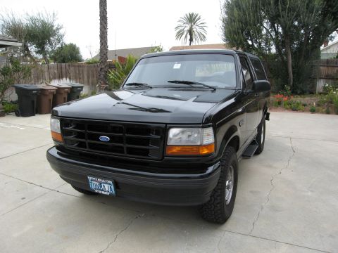 1986 Ford Bronco - A black 1990s Ford Bronco is parked on a concrete driveway in front of a wooden fence and some greenery. The front of the vehicle is angled…