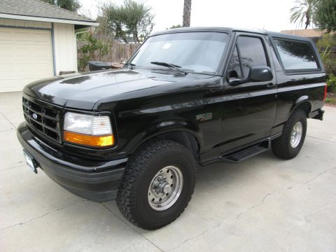 1986 Ford Bronco - A black 1980s Ford Bronco is parked on a concrete driveway in front of a garage. The vehicle is shown from a 3/4 front angle, highlighting…