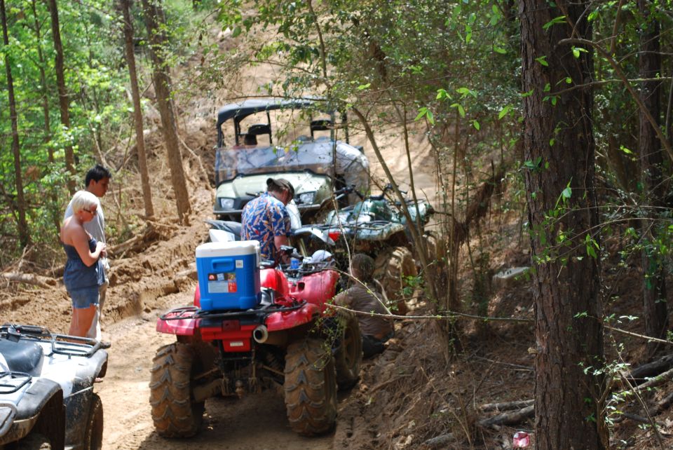 Muddy trail with several ATVs and people. A red ATV with a blue cooler is in the foreground.