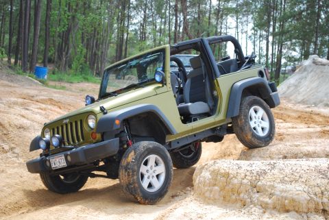 2008 Jeep Wrangler - A green Jeep Wrangler is shown off-roading on a dirt track with large rocks. The vehicle is angled, with its front wheels on a higher eleva…