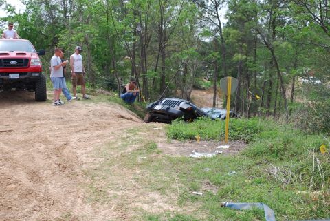 2008 Jeep Wrangler - A black Jeep Wrangler is tipped on its side in a ditch next to a dirt path. Several people are standing around a red Ford pickup truck in t…