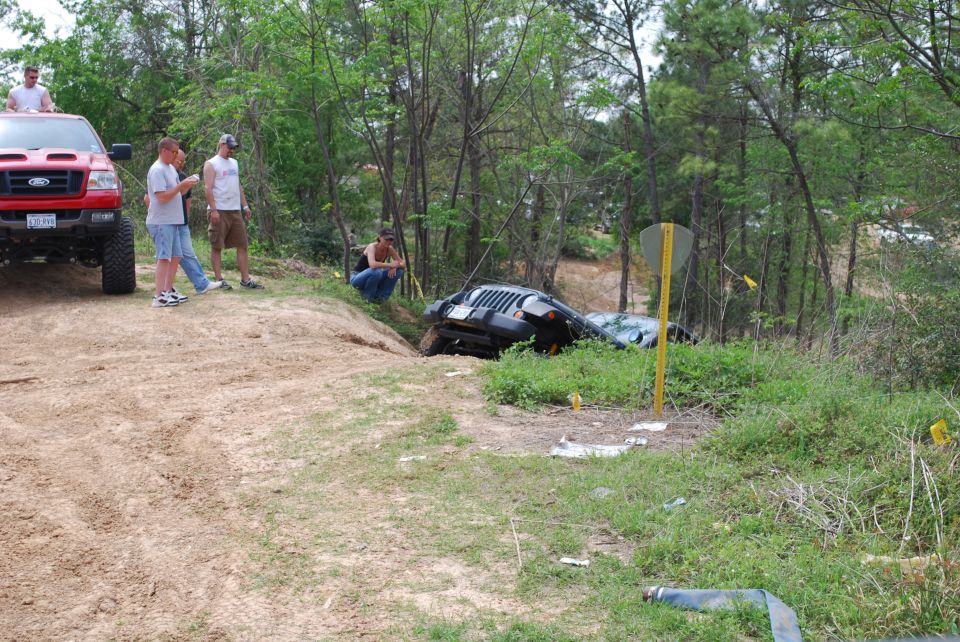 Black Jeep Wrangler tipped on its side in a ditch, with people near a red Ford pickup truck.
