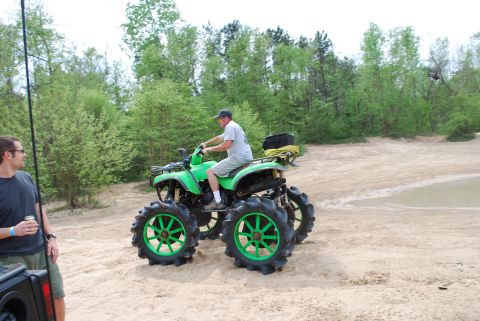 2008 Jeep Wrangler - A man rides a bright green ATV with oversized tires on a sandy track surrounded by trees. Another man stands to the left, holding a drink.