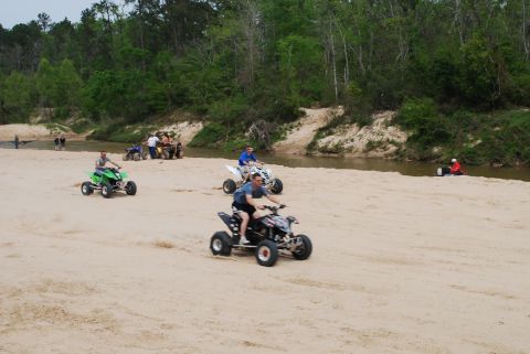 2008 Jeep Wrangler - Multiple people are riding ATVs on a sandy riverbank with trees in the background. One ATV is green and being ridden by a man in a grey shi…