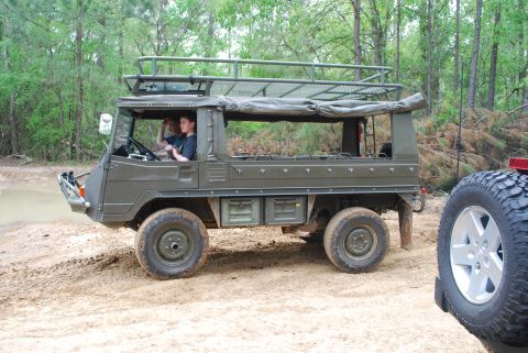2008 Jeep Wrangler - A vintage olive green all-terrain vehicle with a roof rack is parked on a dirt path next to a body of water. Two young people are visible i…