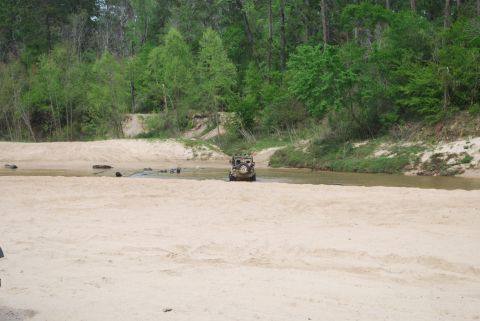 2008 Jeep Wrangler - A tan Jeep Wrangler is driving through a shallow, sandy riverbed. The vehicle is seen from the rear, with lush green trees lining the banks…