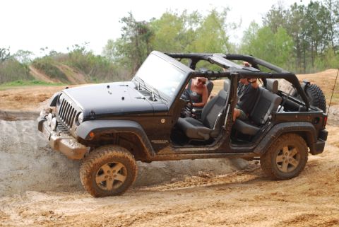 2008 Jeep Wrangler - A black Jeep Wrangler is shown from the side, driving on a dirt path with some mud. The doors are off, and two people are visible inside.