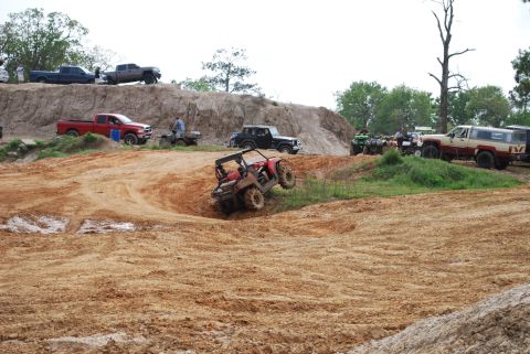 2008 Jeep Wrangler - A red UTV is shown at an extreme angle, appearing to be stuck or performing a stunt in a muddy off-road park. Several other vehicles, inclu…