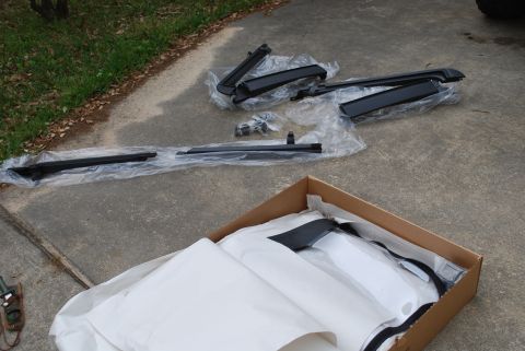 2008 Jeep Wrangler - Several black automotive roof rack components are laid out on a concrete surface, some still in plastic packaging. A cardboard box containi…