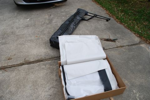 2008 Jeep Wrangler - A new black soft top kit laid out on a concrete driveway, with the folded vinyl top and bow frame beside an open cardboard box holding tiss…
