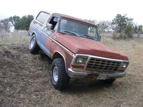 1979 Ford Bronco - A weathered, two-tone red and blue Ford Bronco is parked on a grassy, uneven terrain with trees in the background. The vehicle is angled to…
