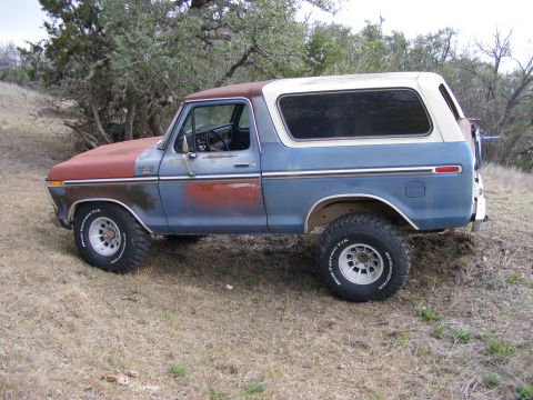 1979 Ford Bronco - The side view of a vintage blue and red Ford Bronco is parked on a dry, grassy hill with trees in the background. The vehicle has off-road…