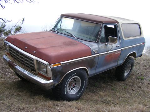 1979 Ford Bronco - A weathered, two-tone 1979 Ford Bronco with a red hood and a blue body sits parked on a grassy incline. The SUV features chrome bumpers, a…
