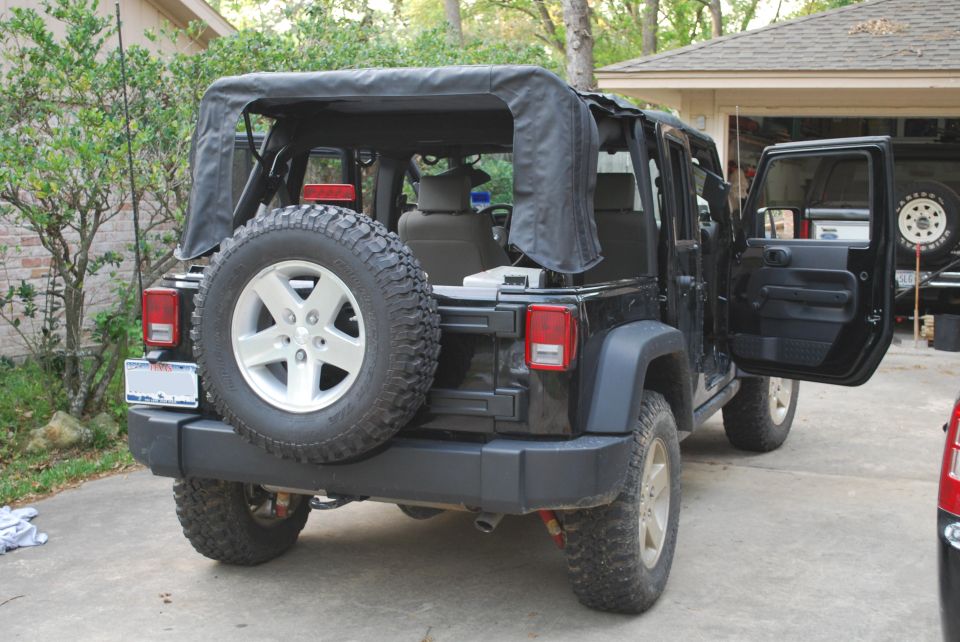 Black Jeep Wrangler rear driver's side view, soft top up, driver door open, spare tire on tailgate.