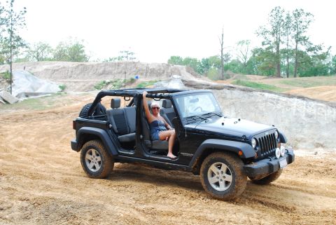 2008 Jeep Wrangler - A woman sits in the driver's seat of a black Jeep Wrangler with the doors and top removed, parked on a dirt track with sandy hills in the b…