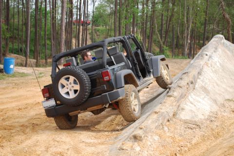 2008 Jeep Wrangler - A black Jeep Wrangler is shown from the rear-side angle as it climbs an obstacle made of dirt and logs. The driver is visible inside the op…