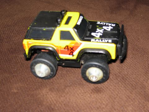 1986 Ford Bronco - A yellow and black toy Ford Bronco rally car is shown from a 3/4 front angle on a brown textured surface. The toy features "4x4 RALLYE" tex…