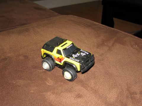 1986 Ford Bronco - A toy 1986 Ford Bronco 4x4 Rallye truck is shown from a 3/4 front angle, resting on a brown textured surface. The toy is yellow with black…
