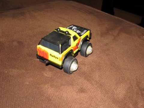 1986 Ford Bronco - A yellow toy 4x4 rally truck with black accents is shown from a rear 3/4 angle, resting on a brown textured surface.