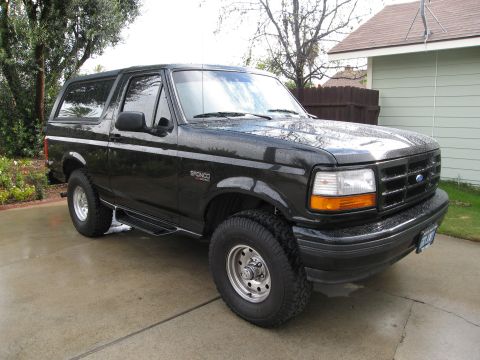1986 Ford Bronco - The front three-quarter view of a black Ford Bronco is shown parked on a wet concrete driveway. The vehicle has chrome wheels and running b…