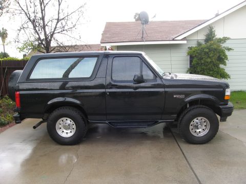 1986 Ford Bronco - A black 1980s Ford Bronco is parked on a wet concrete driveway in front of a house. The vehicle has a spare tire mounted on the rear and af…