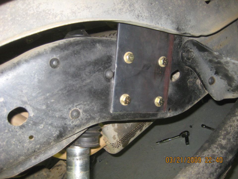 Close-up of a metal bracket bolted to a vehicle's frame near the suspension. A wrench is on the ground