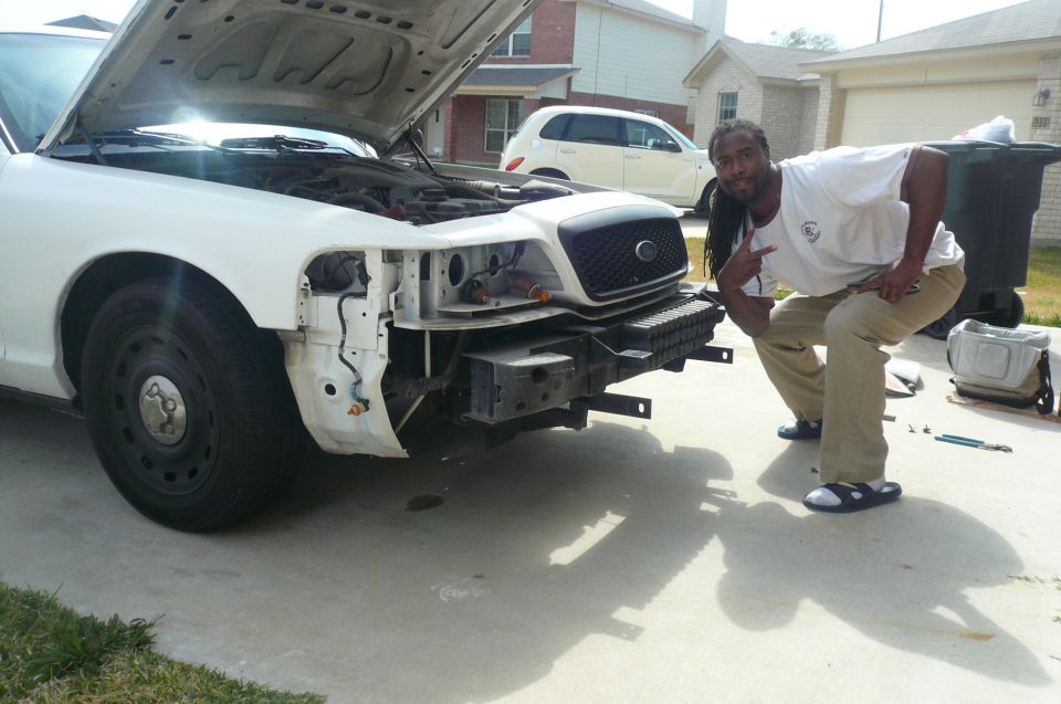 White Crown Victoria with hood up and front bumper removed, man posing beside it in driveway