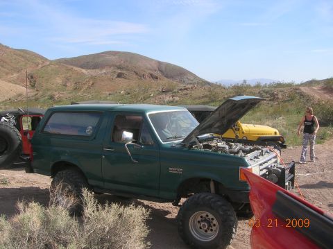 1985 Ford Bronco - A green Ford Bronco is parked in a desert area with its hood up, next to a yellow Jeep. A woman stands nearby.