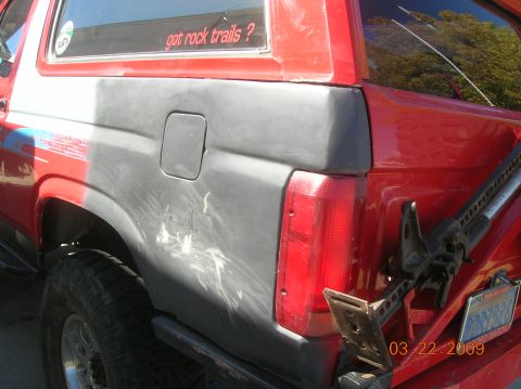 1985 Ford Bronco - Rear view of a red SUV with a gray primer-covered quarter panel and a Hi-Lift jack attached to the tailgate.