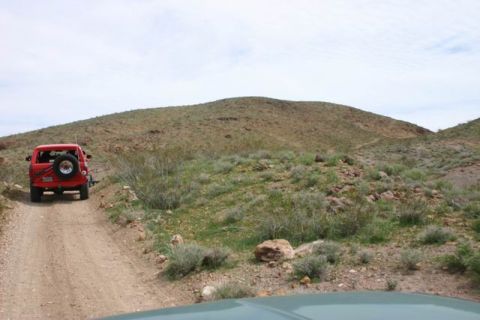1991 Ford Bronco - A red SUV drives along a dusty trail through a barren, hilly terrain under a cloudy sky.