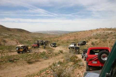1991 Ford Bronco - A convoy of off-road vehicles navigates a dusty trail through a desert terrain under a partly cloudy sky.