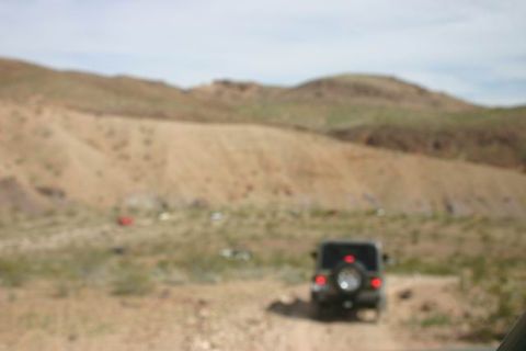 1991 Ford Bronco - A black SUV is parked on a dirt road in a desert landscape with hills in the background. Other vehicles are visible in the distance.
