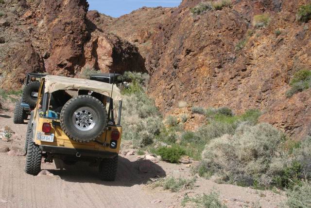 Yellow Jeep Wrangler with tan soft top, spare tire on rear, driving on dirt road