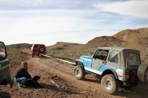 1991 Ford Bronco - A blue Jeep is being towed by a red Jeep on a dusty desert trail, with a woman and her dog observing the scene.