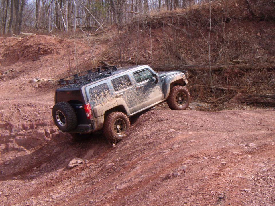 Mud-covered silver Hummer H3 SUV with a roof rack and spare tire on a dirt trail